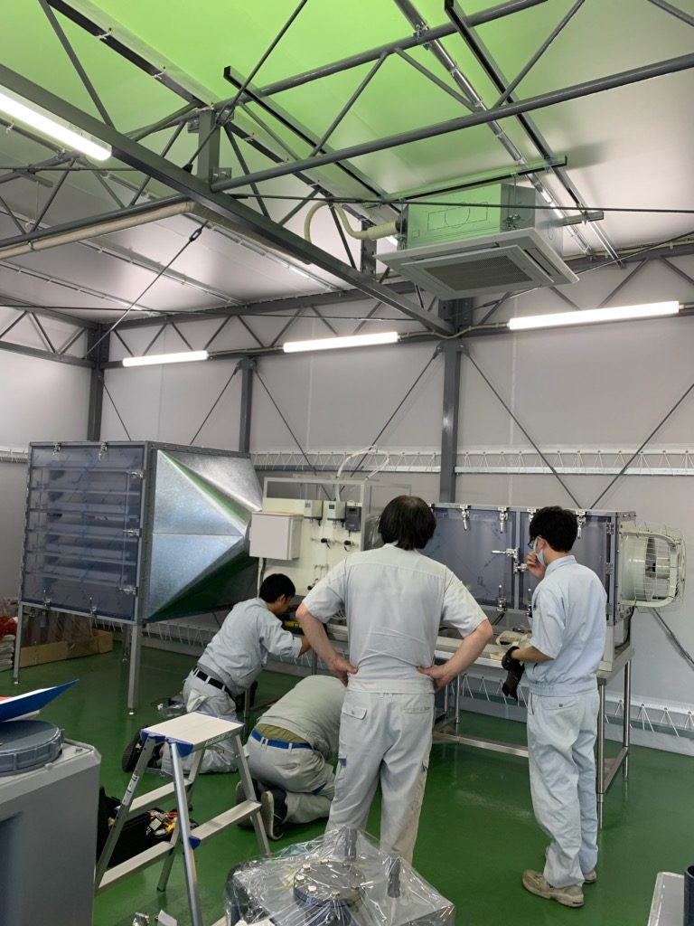 断熱パネルで囲われた試験室に据えられた、送風機・ダクト・箱型ユニットからなる大型の試験装置。作業着姿の数人が組み立てを進めている。